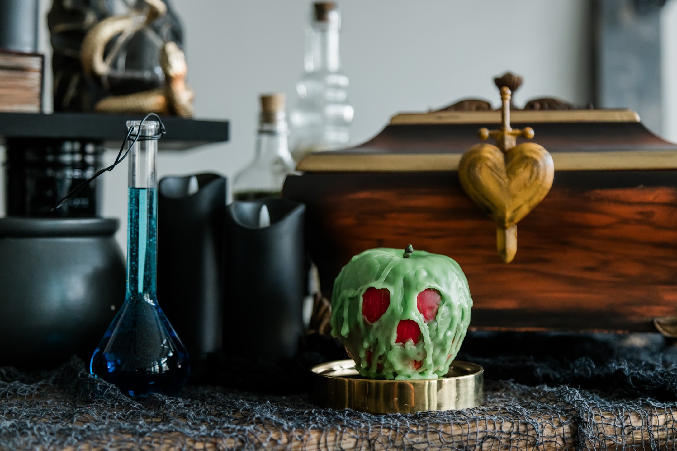 A spooky setup features a green poison apple with red eyes and mouth on a gold plate, a blue potion in a flask, black goblets, and a wooden chest adorned with a gold heart. The scene is set on a netted fabric, suggesting a Halloween or witchcraft theme.